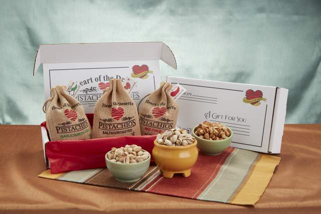 Open gift box from Heart of the Desert, featuring bags of pistachios including Garlic and Green Chile, Salted, and Red Chile flavors, along with three bowls of shelled pistachios on a cloth-covered table.