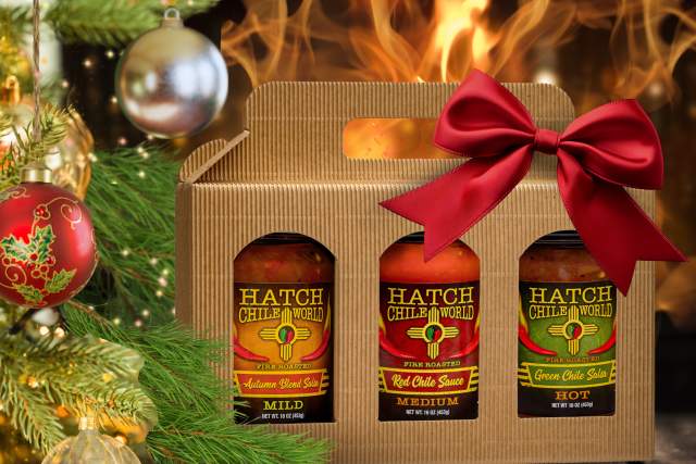 Gift box of Hatch Chile World sauces with a red bow, nestled beside Christmas decorations and a cozy fireplace.