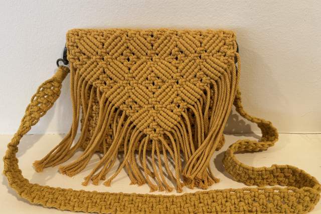 A yellow crocheted bag with fringe detail sits in front of white wall on a white table.