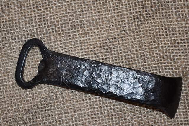 A rustic, hand-forged bottle opener with a textured metal finish lies on a burlap surface.