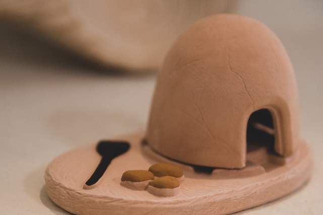 Miniature clay model of an adobe oven (horno) with a wooden paddle and small round bread loaves on a base.
