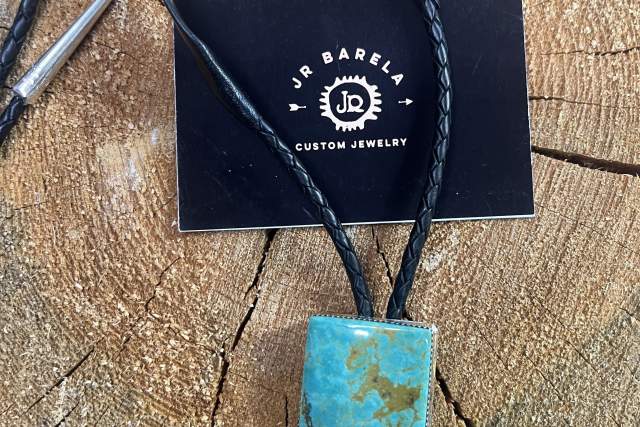 Jewelry piece with a turquoise stone pendant displayed on a braided leather cord. The pendant rests on a wooden surface alongside a business card for JR Barela Custom Jewelry.