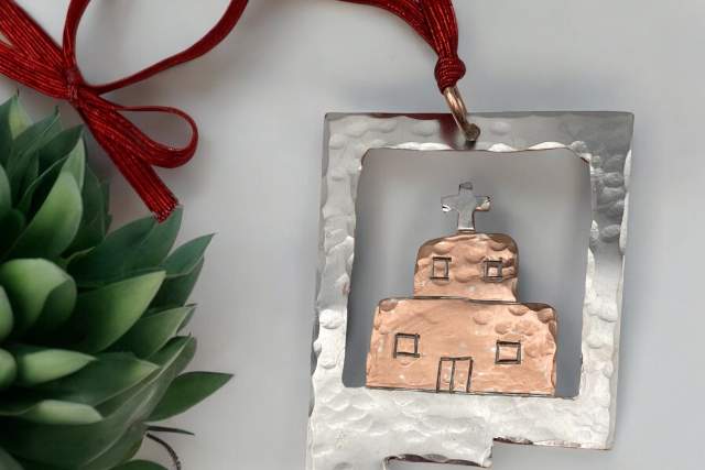 A decorative metal ornament depicting a church, with a cross atop the roof, hangs by a red ribbon beside a green succulent plant on a light background.