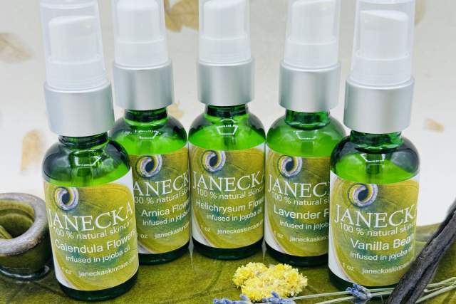 Six bottles of Janécka Jojoba oils, including Calendula Flower, Arnica, Helichrysum, Lavender, and Vanilla Bean, arranged on a marble surface with scattered dried flowers and leaves.