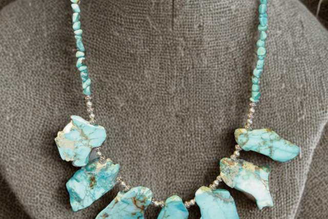 A necklace displayed on a burlap-covered mannequin bust. The necklace features irregular turquoise stones interspersed with small silver beads.