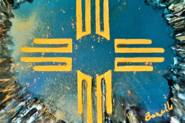 Abstract painting on a coaster featuring a central symbol in yellow, resembling the Zia sun symbol from the flag of New Mexico, set against a textured dark blue and black background. Signed by the artist 'Brad' in the lower right corner.