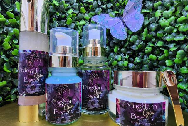 A display of Kingdom of Beauty skincare products, including a lavender-toned serum, moisturizer, and a clear gel product, arranged in front of a green leafy background with a decorative blue butterfly.