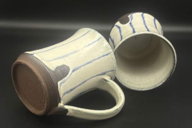 Two handcrafted stoneware mugs with blue stripes and tan bases are lying on their sides on a dark surface.