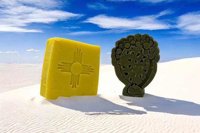 Two bars of soap, one shaped like the symbols from the flags of New Mexico and one shaped like a bundle of herbs, placed on a sandy surface under a clear blue sky.