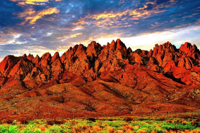 Sunset over the rugged terrain of the Organ Mountains-Desert Peaks National Monument, highlighting vibrant red rocks and a sky streaked with orange and blue.