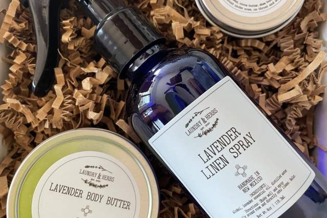 An array of Lavender & Birch beauty products including Lavender Linen Spray in a spray bottle, Lavender Body Butter, and Dream Balm, displayed on crinkled paper filling inside a box.