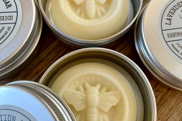Several open tins of lotion bars, each embossed with a bee design, displaying labels that include "Lavender" and "Handmade in New Mexico."