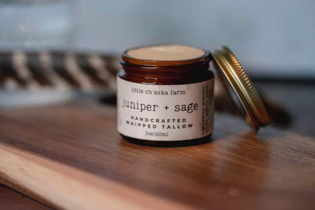 A small jar of "Little Ch'aska Farm Juniper + Sage Handcrafted Whipped Tallow" on a wooden surface with its lid placed beside it.