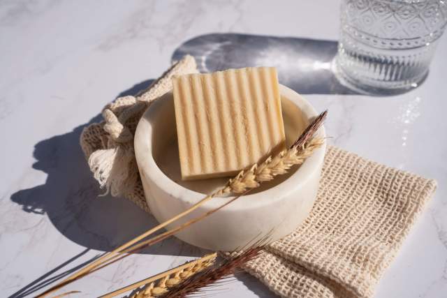 A bowl of rectangular oat biscuits with a wheat stalk on a textured napkin, set on a marble surface beside a glass of water.