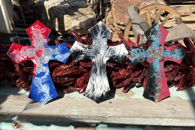 Three decorative crosses, each uniquely painted in red, black, and turquoise patterns, standing upright on a bed of chili peppers on a wooden surface.