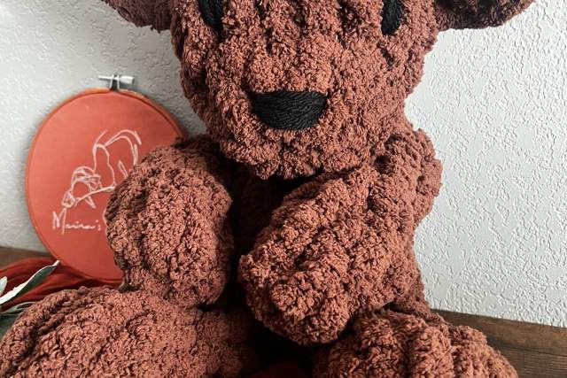 A plushie of a brown bear sits against a white wall.