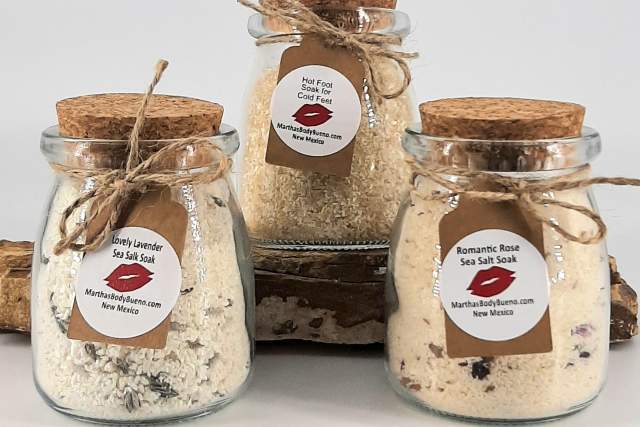 Three glass jars filled with bath soaks are displayed. Each jar has a cork lid and a label tied with twine. They are arranged on a rustic wooden block.