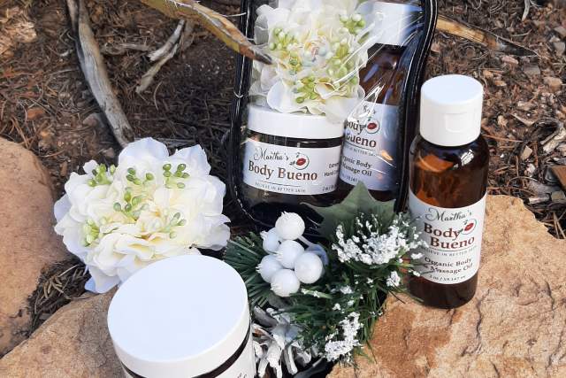 A display of Body Bueno skincare products arranged on a rock, among natural surroundings. The products include jars and bottles with visible labels, accompanied by white flowers.