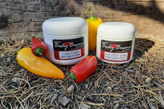 Two Martha's Body Bueno brand soak products on a bed of straw, with a yellow and a red pepper alongside. The left container is labeled "I Like It Hot Sea Salt Soak" and the right one is "Hot Foot Soak for Cold Feet.