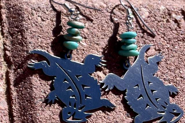A pair of earrings featuring dark silhouettes of horned toads, decorated with turquoise beads, displayed against a red brick background.