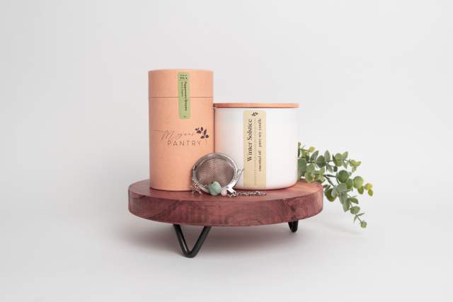 Two "Megan's Pantry" branded candles on a wooden shelf with a small plant and a silver loose tea strainer.