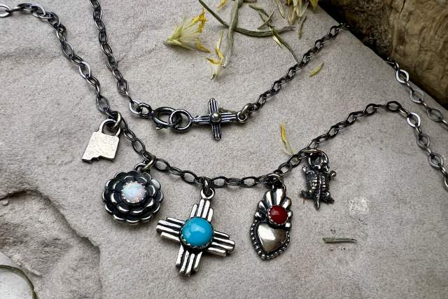 A collection of Southwest-style silver jewelry featuring a necklace with pendants including a flower, a turquoise gem, and a red stone, arranged on a rocky surface with scattered yellow petals.
