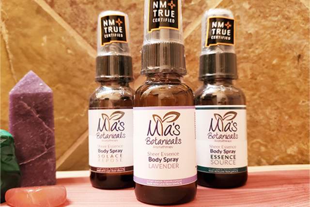 Three bottles of Mia's Botanicals body spray with lavender scent, displayed in front of a wooden background, flanked by colorful crystals.