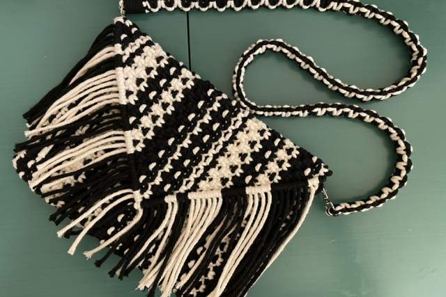 A handmade, triangular macramé bag with woven patterns and fringe detailing. The bag has an attached braided strap in matching colors, resting on a flat surface.