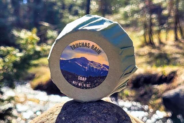A package of Goat Milk Soap sits on a rock before a winding stream in the midst of a green forest. Packge reads Truchas Rain Soap, Net Weight 4oz