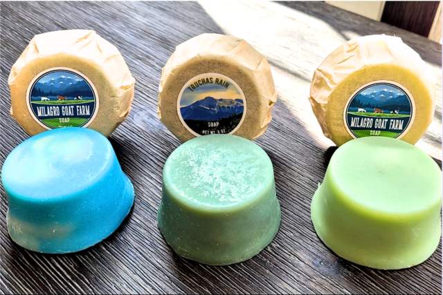 Three bars of soap in front of three packages with the label "Milagro Goat Farm" on a wooden surface. The soap colors are blue, green, and pale green.