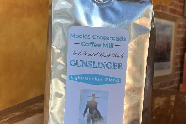 A sealed bag of Mock's Crossroads Coffee Mill's Gunslinger Light-Medium Blend coffee, standing on a wooden table against a brick wall background. The label features the name and location, Clayton, New Mexico.