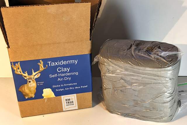 A product display of Air-dry Taxidermy Clay, marketed as sticking to anything without cracking. The packaging box, with an image of a deer head sculpture and text, is slightly open revealing the grey clay wrapped in plastic beside it. Additional text on the image highlights the clay's compatibility with all kinds of paint.