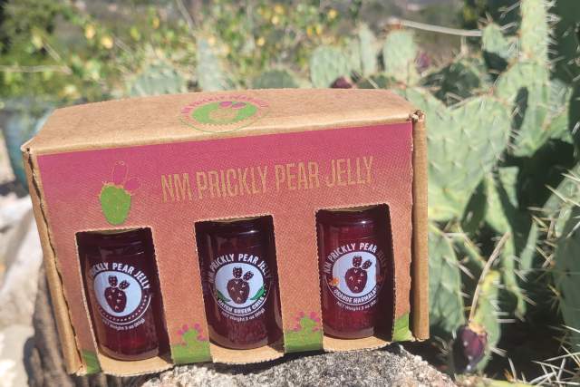 A pack of mini prickly pear jelly jars from Santa Cruz Chili & Spice Company, placed on a rocky surface, with a cactus plant and clear blue sky in the background.