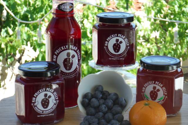 Assorted jars of "NM Prickly Pear Jelly" products, including syrup and marmalade, displayed on a table with a bowl of blueberries and an orange, with greenery in the background.
