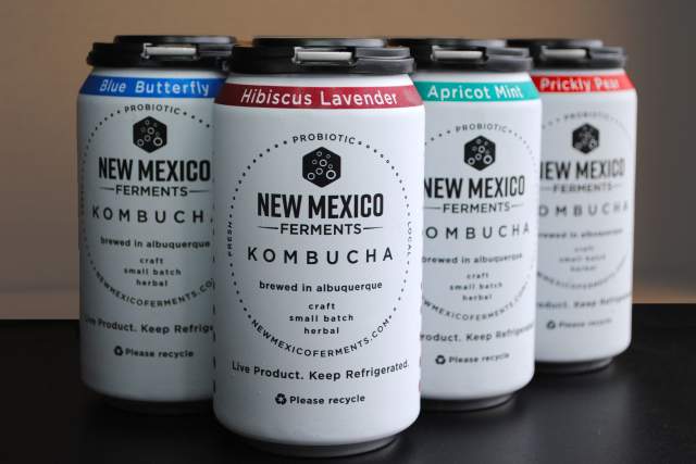 Three cans of New Mexico Ferments kombucha on a table, featuring flavors Hibiscus Lavender, Apricot Mint, and Prickly Pear. Each can is blue with white and black text detailing the brand and flavor.