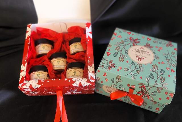 Gift box containing three jar candles, tied with a red ribbon, next to a closed box with a festive design, all on a black background.