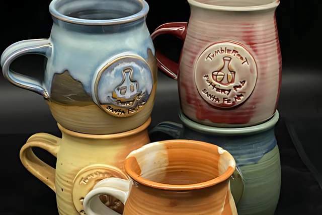 Four ceramic mugs of varying colors and designs, featuring distinct emblems and glazed finishes. Two mugs display logos: one with a "Santa Fe" Inscription and one with a "Tumbleroot" inscription