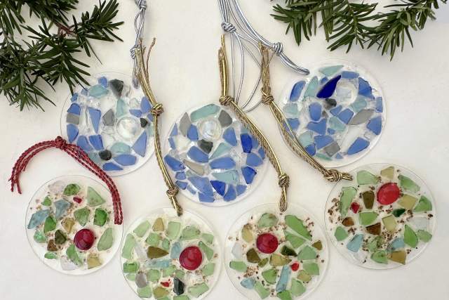 Handmade circular mosaic ornaments in various colors, including blue and green shades, displayed on a white surface with evergreen sprigs in the background