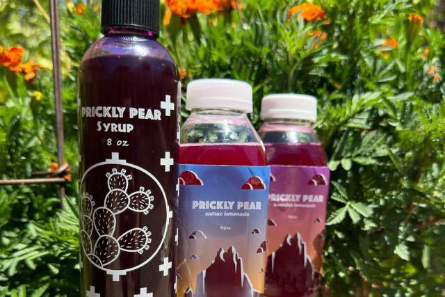 Three bottles of prickly pear syrup are displayed against a backdrop of vibrant orange flowers and green foliage. From left to right: an 8 oz dark purple bottle labeled "Prickly Pear Syrup," and two smaller translucent bottles containing pinkish-red prickly pear syrup, both labeled with the same name.
