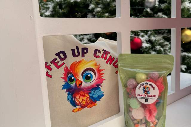 A tote bag with an illustrated owl and the phrase "Puffed Up Candy" next to a package of Candy Salad gummies, displayed in front of a frosted window with a view of a decorated Christmas tree.