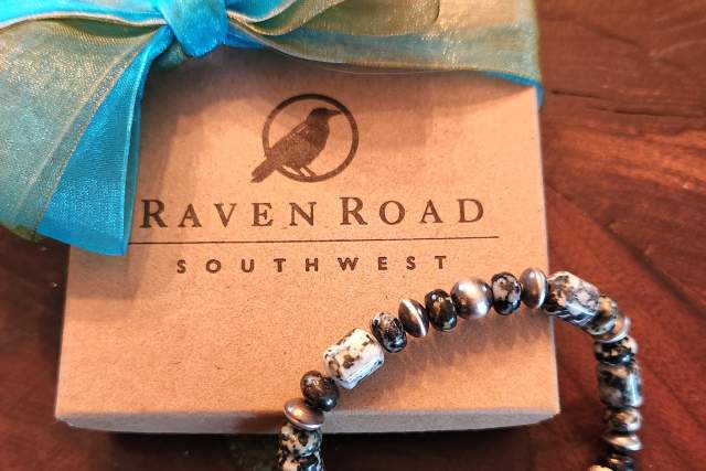 A decorative bracelet with various beads rests in front of a gift box adorned with a teal ribbon, featuring the logo "Raven Road South West.