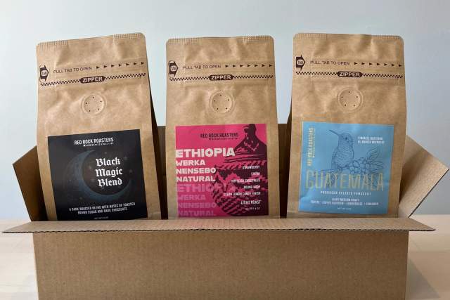 Three bags of Red Rooster Coffee standing upright in a cardboard box on a table.