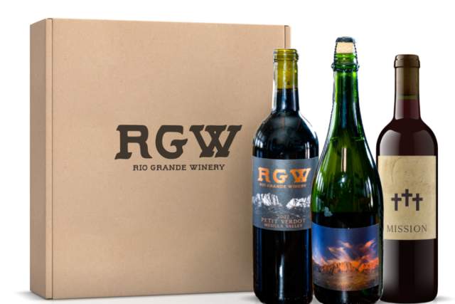 A cardboard box labeled "RGW Rio Grande Winery" next to three bottles of wine with labels that include "Petit Verdot" and "Mission," featuring scenic imagery on the label.
