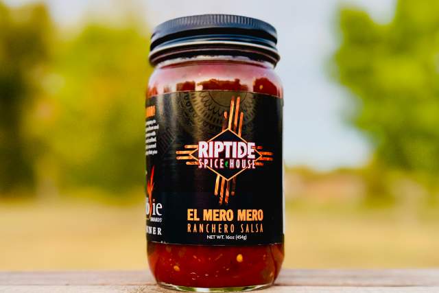 A jar of Riptide Spice House's "El Mero Mero Ranchero Salsa" on a wooden surface with a blurred natural background.