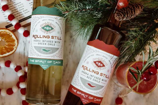 Two bottles of Rolling Still vodka, one infused with green chile and the other with red chile, surrounded by festive holiday decorations including pine branches and sliced oranges.