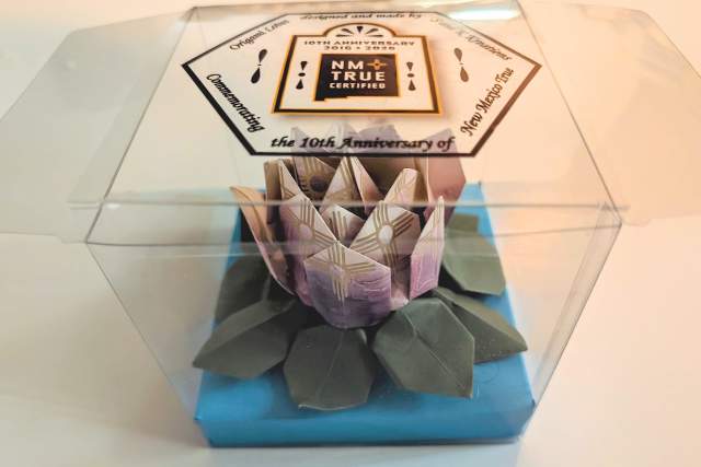 This image shows a decorative item resembling a flower with geometric patterns inside a clear plastic box. The box is labeled with "NM TRUE CERTIFIED" and mentions the 10th anniversary of New Mexico True.