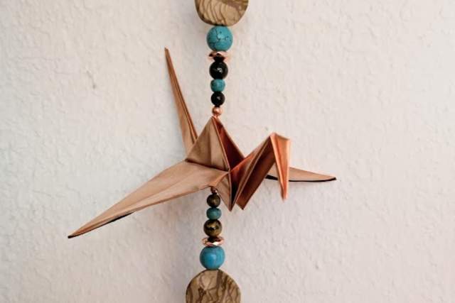 A handmade decorative piece featuring two origami cranes and strung beads, with an affixed label stating "Created by K KReations Handmade with Love." The decoration is suspended from a metal ring, and it includes natural and teal beads interspersed with metallic elements and a spiral pendant at the bottom.
