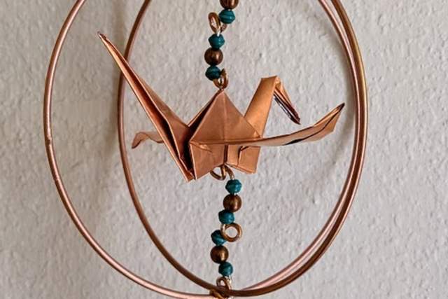 A copper-colored origami crane suspended within a circular frame, adorned with beads, and attached to a cone-shaped pendant, hanging against a textured white wall.