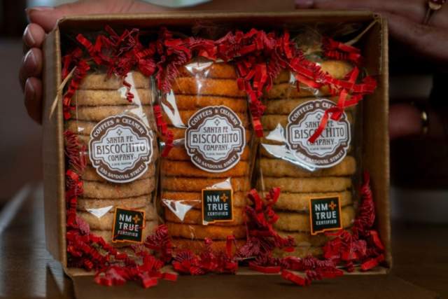 A person holding a gift box from Santa Fe Biscochito Company, containing three clear packages of traditional cookies, each tied with red ribbons, labeled barer and biscochitos.
