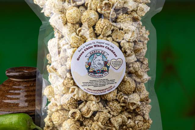 A clear bag of popcorn is displayed. The background includes green and orange hues, with various ingredients like cheese, green peppers, and seeds surrounding the bag.